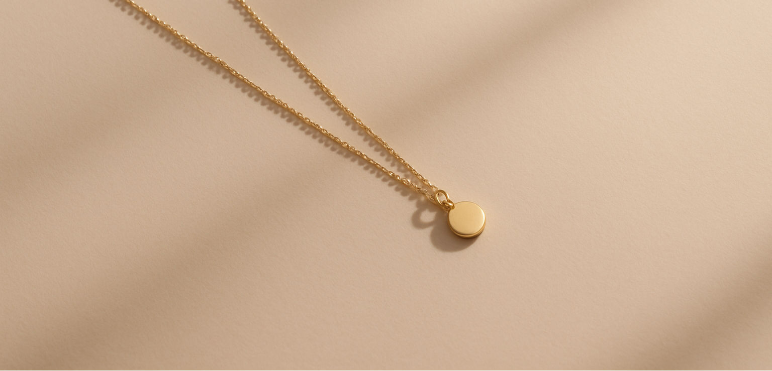 Minimalist editorial hero image of jewelry — a single delicate gold necklace with a small pendant placed diagonally on a soft beige background, with elegant soft shadows and lots of negative space. The composition should feel refined, luxurious and calm, with a modern aesthetic. No clutter, no text, no people.